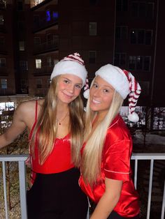 two girls in santa hats posing for the camera on a balcony at night time with snow and buildings behind them