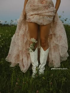 a woman wearing white cowboy boots standing in the middle of a field with flowers on her dress