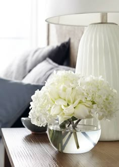 a vase with white flowers sitting on a table next to a lamp and bed in the background