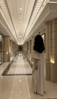 a woman in a long white dress is walking down the hall with her back to the camera