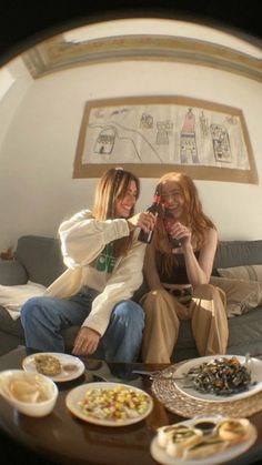 two women sitting on a couch with plates of food in front of them, looking at the camera through a fish eye lens