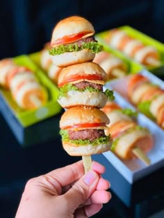 a hand holding up a mini hamburger on a stick in front of trays of food