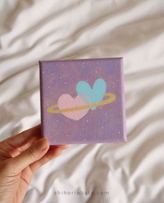 a hand holding up a small purple box with hearts and saturn in the sky on it