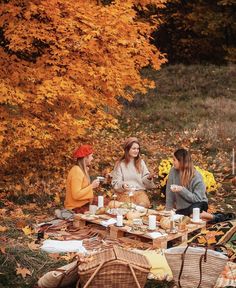 three women sitting at a picnic table with food and drinks in front of trees that have yellow leaves on them