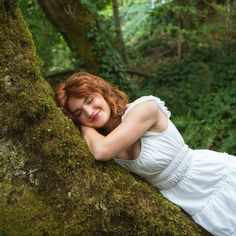 a woman in a white dress is leaning on a mossy tree and smiling at the camera