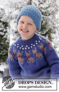 a young boy in a blue sweater and hat smiles at the camera while standing in front of snow covered trees