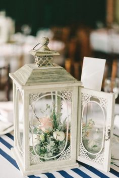 a small white lantern with flowers inside on a striped table cloth covered place setting at a wedding reception