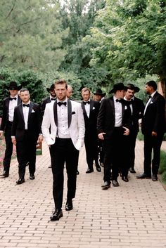 a group of men in tuxedos walking down a brick path together with trees and bushes behind them