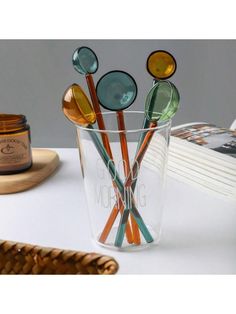 a glass vase filled with lots of spoons on top of a white table next to a book