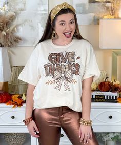 The Give Thanks With Bow Graphic Shirt is a charming and stylish choice for Thanksgiving celebrations. This shirt is available in a natural color and comes in sizes YXS-5XL, offering options for everyone. With the uplifting phrase "Give Thanks" adorned with a cute bow design, it embodies the spirit of gratitude and family gatherings. You can choose between short sleeve or long sleeve styles, making it suitable for any fall weather. Whether you're hosting a dinner or enjoying a cozy family gather Beige Short Sleeve Tops For Fall, Beige Crew Neck T-shirt For Fall, Casual Beige T-shirt For Fall, Beige Graphic Print T-shirt For Fall, Beige Short Sleeve Shirt For Fall, Beige Relaxed Fit T-shirt For Fall, Relaxed Fit Beige T-shirt For Fall, Bow Graphic, Uplifting Phrases