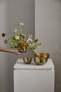 two gold bowls holding flowers on top of a white pedestal in front of a gray wall