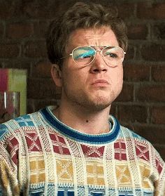 a man wearing glasses standing in front of a brick wall