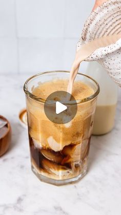 a person pouring milk into a glass filled with ice and brown liquid on top of a marble counter