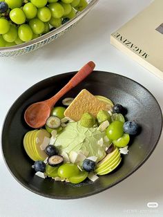 a black bowl filled with fruit and cheese next to a book on a white table