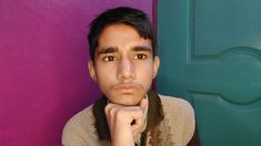 a young man leaning against a purple wall with his hand on his chin and looking at the camera