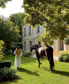 a woman walking with a horse in front of a large house on a sunny day