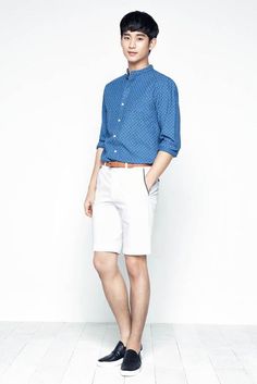 a young man standing in front of a white wall wearing shorts and a blue shirt