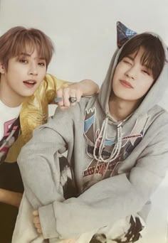 two young men sitting next to each other in front of a white wall wearing hoodies