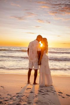 a man and woman kissing on the beach at sunset, with footprints in the sand