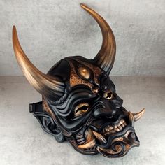 a black mask with gold accents and horns on it's head is shown in front of a gray background