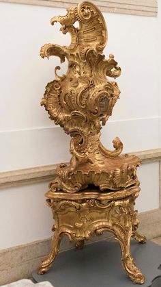 an ornately decorated golden chair sitting on top of a gray table next to a white wall