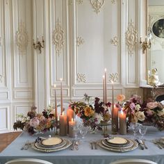 the table is set with candles, plates and flowers in front of an ornate wall