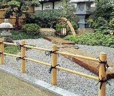 a wooden fence in front of a building with trees and bushes around it, along with rocks and stones on the ground