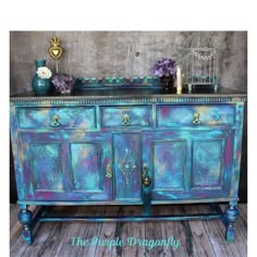 an old dresser painted in blue and purple with gold accents on the top, sitting on a wooden floor next to a birdcage