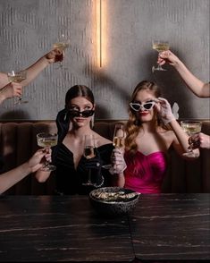 three women wearing sunglasses and holding wine glasses in front of their faces while sitting at a table