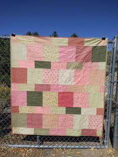 a pink and green patchwork quilt hanging on a chain link fence in front of some trees