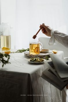 a person holding a wooden stick over a cup of tea on a table next to a book