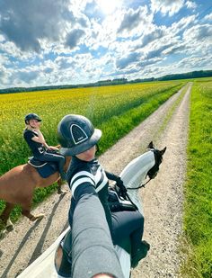 two people riding horses on a dirt road next to a field with grass and yellow flowers