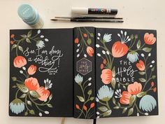 two notebooks with flowers on them next to markers and pens, one has the bible written in it