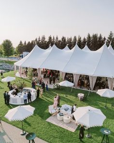 a group of people standing around tables and umbrellas in the grass near some trees