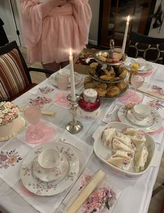 a table topped with plates and cakes next to a woman in a pink dress holding a lit candle