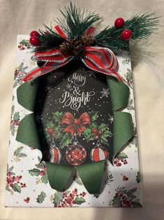 a christmas card with holly wreaths and bells on it, hanging from a ribbon