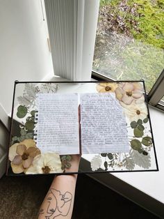 a person holding up a book with flowers on it and writing in the pages, next to a window