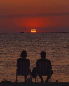 two people sitting in lawn chairs watching the sun go down over the ocean at sunset