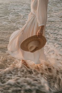 a woman in a white dress and straw hat walking into the ocean