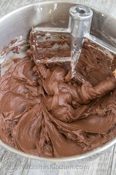 a metal bowl filled with chocolate frosting on top of a wooden table next to a spatula