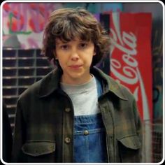 a young boy in overalls standing next to a coca - cola sign and looking at the camera