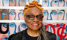 an older woman wearing glasses and a yellow head scarf stands in front of colorful paintings