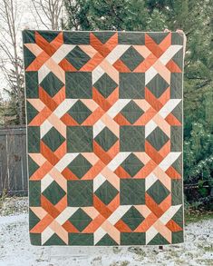 an orange and green quilt is hanging on a fence in front of some snow covered trees