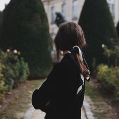 a woman standing in front of a building with trees and bushes behind her back to the camera