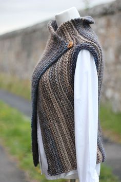 a mannequin wearing a knitted sweater and vest on a wooden stand in front of a stone wall