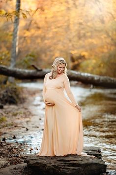 a pregnant woman in a long dress standing on rocks near a river