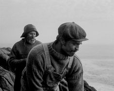 black and white photograph of two men standing on rocks near the ocean, one holding a cell phone