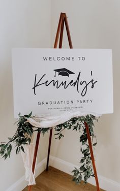 a welcome sign for a graduation party with greenery on the bottom and black lettering that reads, welcome to kenley's graduation party