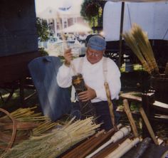 an old man sitting in front of a pile of brooms holding a jar with some sort of liquid