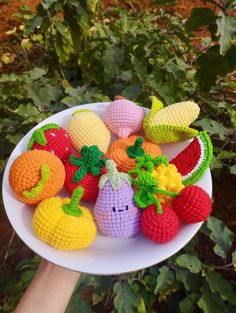 crocheted fruits and vegetables on a white plate in front of some bushes with leaves